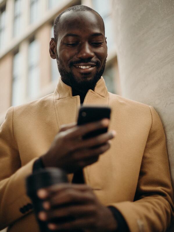 EMAILS - Dark-skinned black man wearing tan jacket and holding cell phone while smiling