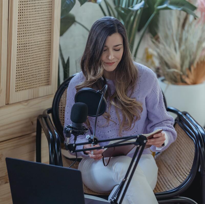 PRODUCTS - 
Light-skinned white woman wearing lavender blouse with white jeans. She has a  podcast microphone in front of her as she looks down. 